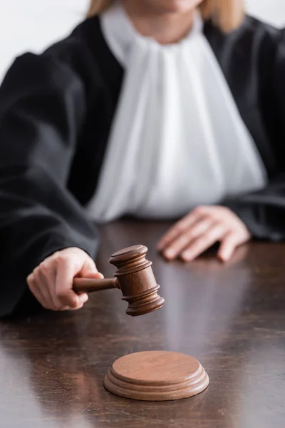 Partial view of judge in black mantle holding wooden gavel on blurred background — Stock Photo
