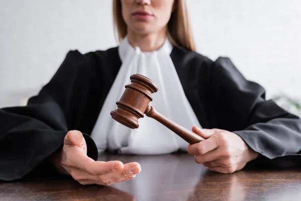 Partial view of judge in black mantle holding gavel while sitting at desk - foto de stock