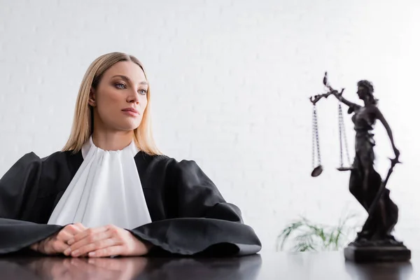 Blonde prosecutor in mantle looking at themis statue on blurred foreground — Photo de stock