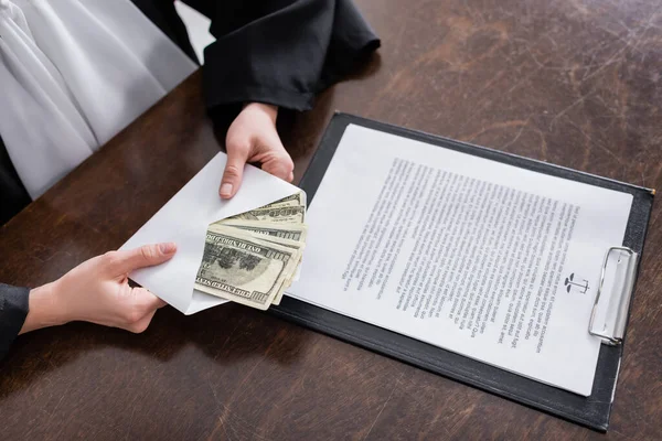 Cropped view of judge holding envelope with dollars near clipboard with lawsuit — Photo de stock