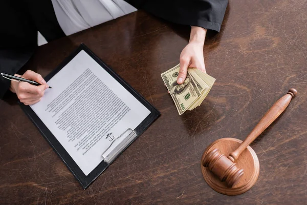 Vista parcial del juez con billetes de dólar firmando veredicto cerca de mazo de madera - foto de stock