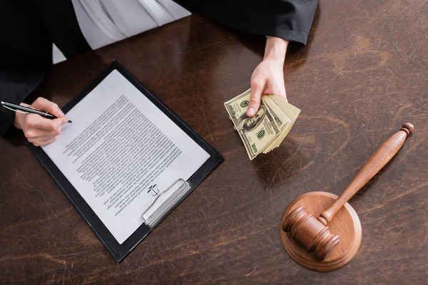 Partial view of judge with dollar banknotes signing verdict near wooden gavel — стоковое фото