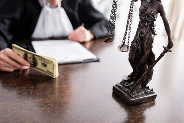 Partial view of blurred judge holding dollars near lawsuit and themis statue - foto de stock