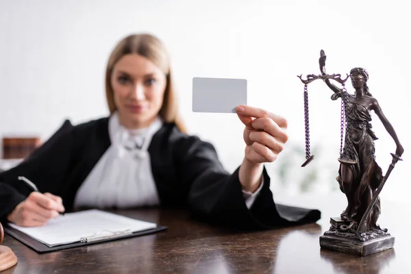 Smiling prosecutor holding empty business card near themis statue, blurred background — Stock Photo