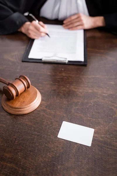 Partial view of blurred judge with pen near wooden gavel and empty business card on desk — Fotografia de Stock