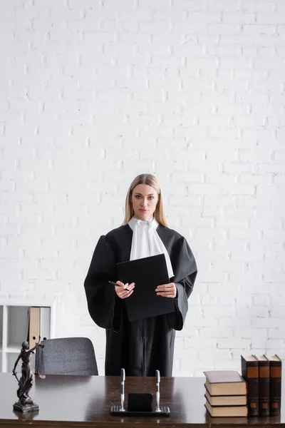 Judge in mantle standing with folder near books and themis statue on work desk — Stock Photo