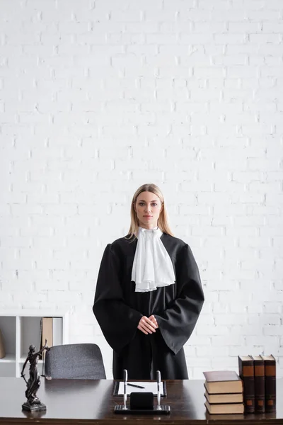 Prosecutor in black mantle looking at camera near desk and white wall in office - foto de stock