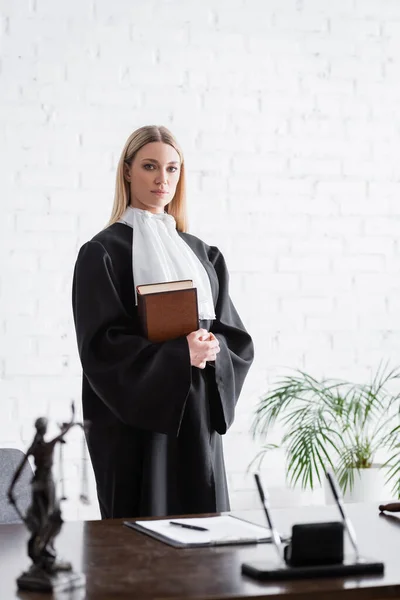 Prosecutor in black mantle looking at camera while standing with book near white wall — Foto stock