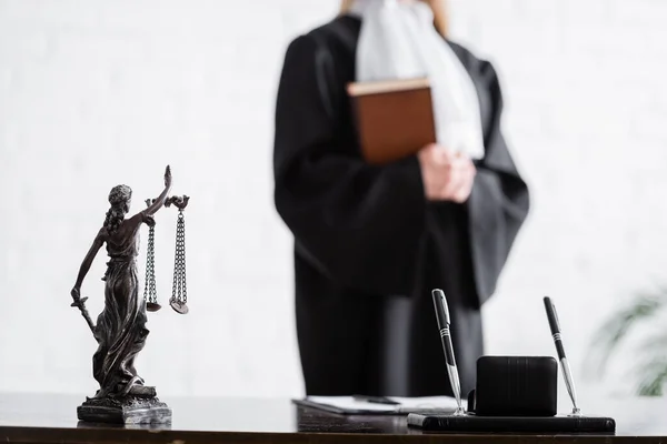 Selective focus of themis statue and pens near cropped prosecutor with book on blurred background — стоковое фото