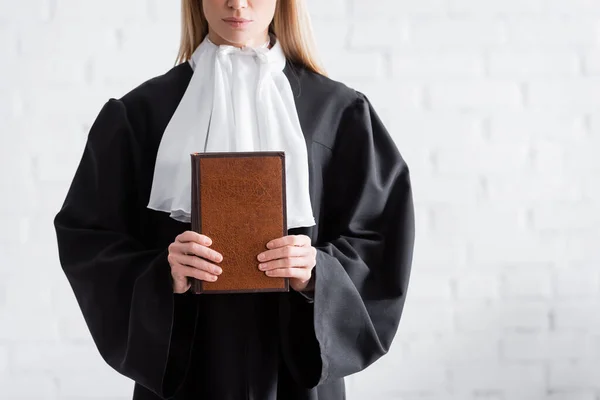 Partial view of prosecutor in black mantle holding book near white wall - foto de stock