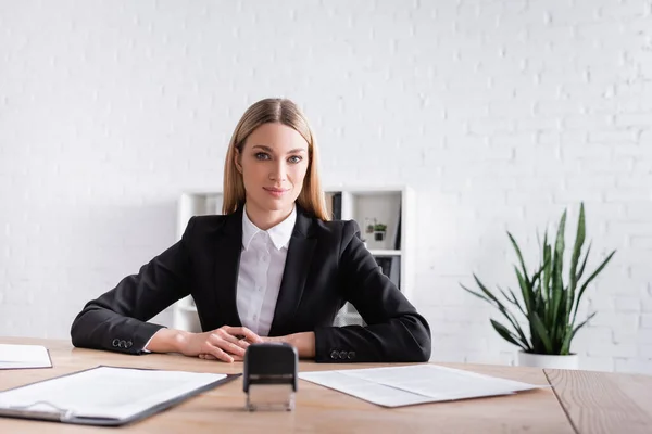 Positive notary looking at camera near documents and stamper on blurred foreground — Stock Photo