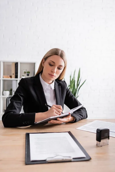 Blonde lawyer writing in notebook near documents and stamper on desk — Stockfoto
