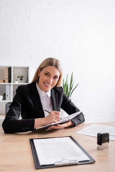 Cheerful notary looking at camera while writing in notebook at workplace — Stockfoto