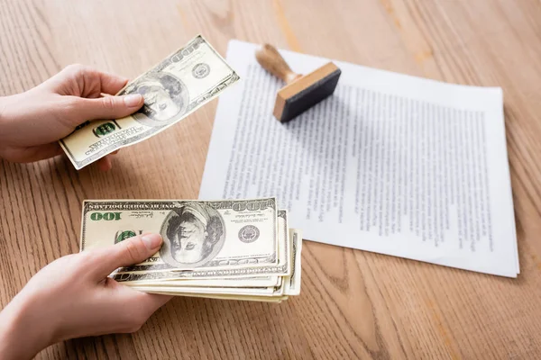 Partial view of lawyer holding currency near stamper and contract on wooden desk — Stock Photo