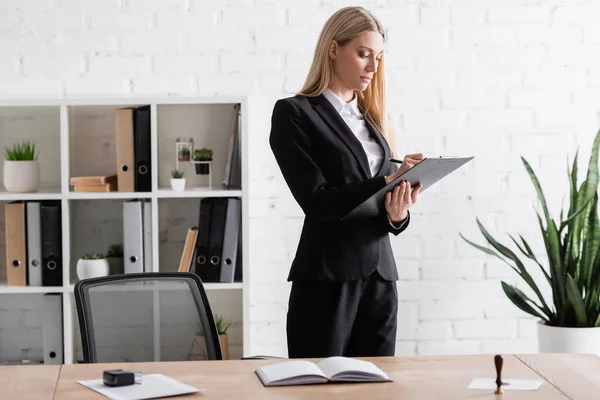 Blonde lawyer writing on clipboard while standing in office - foto de stock