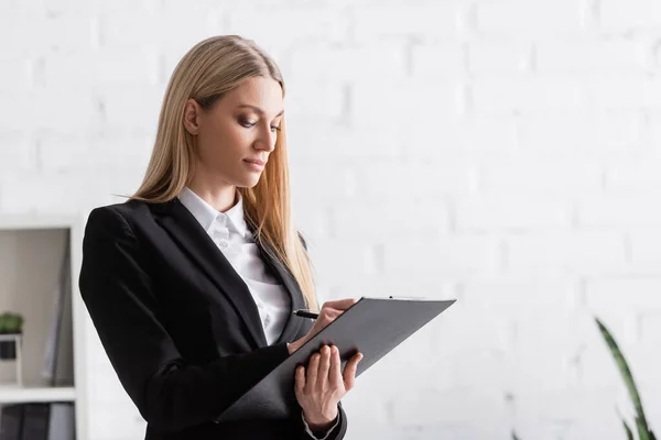 Blonde Anwältin schreibt auf Klemmbrett, während sie in der Nähe einer verschwommenen weißen Wand steht — Stockfoto