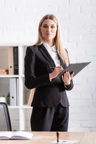 Blonde notary with clipboard and pen looking at camera in office — Foto stock