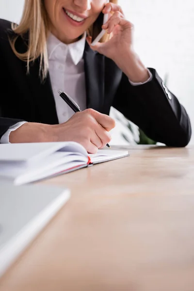 Partial view of smiling notary talking on smartphone and writing in notebook on blurred foreground - foto de stock