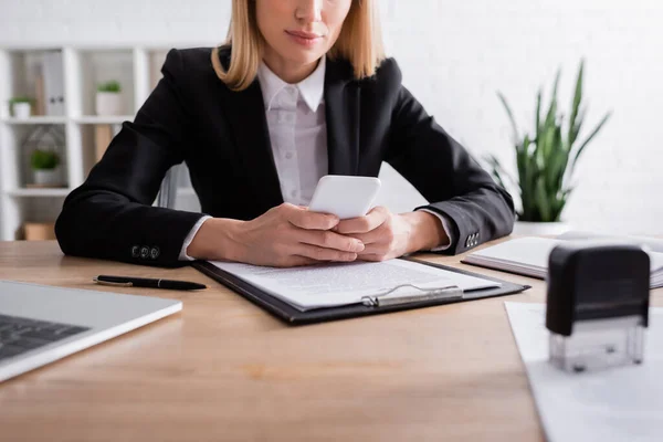 Vista parcial del notario usando smartphone cerca del portapapeles y estampador borroso - foto de stock