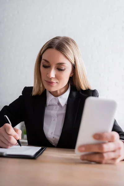 Lawyer holding smartphone and writing in notebook while working in office — Fotografia de Stock