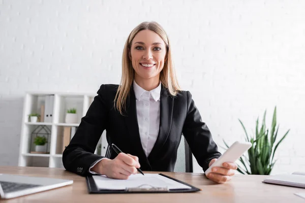 Happy lawyer looking at camera while holding smartphone and pen near clipboard — Stockfoto