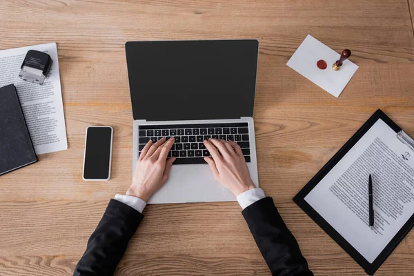 Top view of cropped lawyer typing on laptop near smartphone with blank screen and contract — стоковое фото