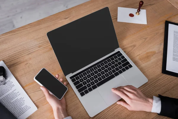 Vista parcial del abogado que sostiene el teléfono móvil cerca del ordenador portátil con la pantalla en blanco y documentos - foto de stock