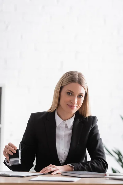 Smiling blonde notary holding stamper and looking at camera while working in office — Photo de stock