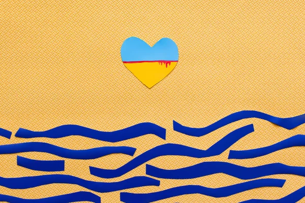 Верхний вид украинского флага с кровью в форме сердца над бумажным морем на текстурированном желтом фоне — стоковое фото