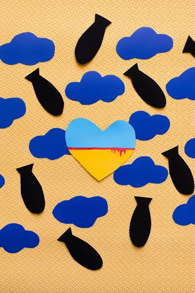 Draufsicht der ukrainischen Flagge in Herzform in der Nähe von Kartonwolken und Papierbomben auf strukturiertem gelben Hintergrund — Stockfoto