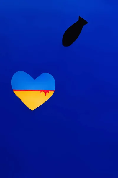 Vista superior de la bomba de papel cerca de la bandera ucraniana con sangre en forma de corazón sobre fondo azul - foto de stock