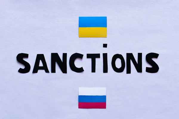 Vue du dessus du lettrage des sanctions près des drapeaux ukrainiens et russes sur fond blanc — Photo de stock