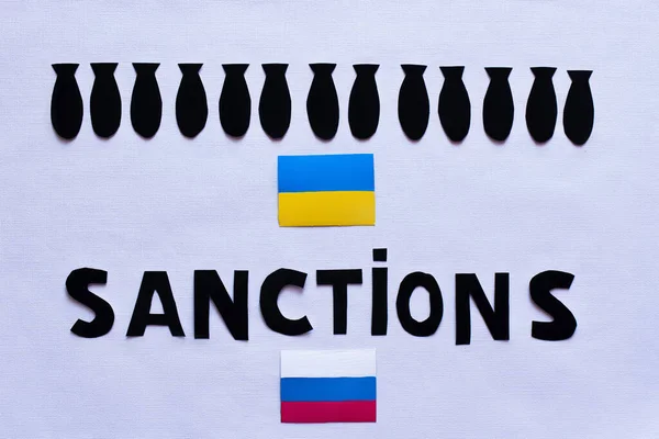 Vue du dessus des bombes à papier, lettres de sanctions près des drapeaux ukrainiens et russes sur fond blanc — Photo de stock