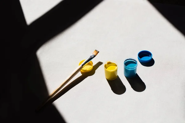 High angle view of blue and yellow paint in jars near paintbrush on white — стоковое фото