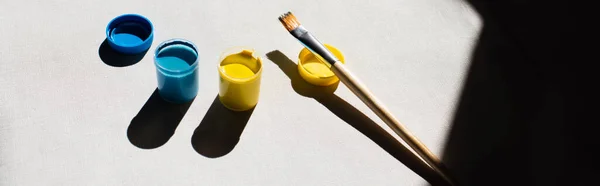 Vista ad alto angolo di vernice blu e gialla in vasi vicino pennello sul bianco, banner — Stock Photo