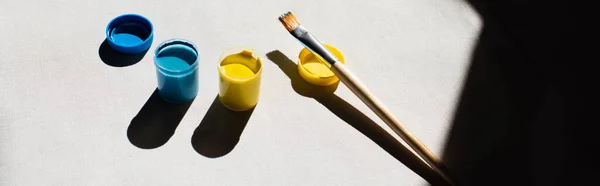 High angle view of blue and yellow paint in jars near paintbrush on white, banner — Stock Photo