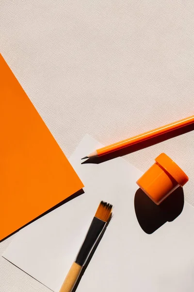 Vue du dessus du pinceau, du crayon et du pot avec de la peinture près du papier orange sur fond texturé blanc — Photo de stock