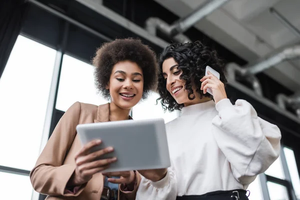 Vue en angle bas de souriantes femmes d'affaires interraciales utilisant une tablette numérique et parlant sur smartphone au bureau — Photo de stock