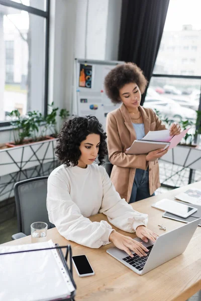 Arabian businesswoman using laptop near african american colleague with papers in office — стокове фото