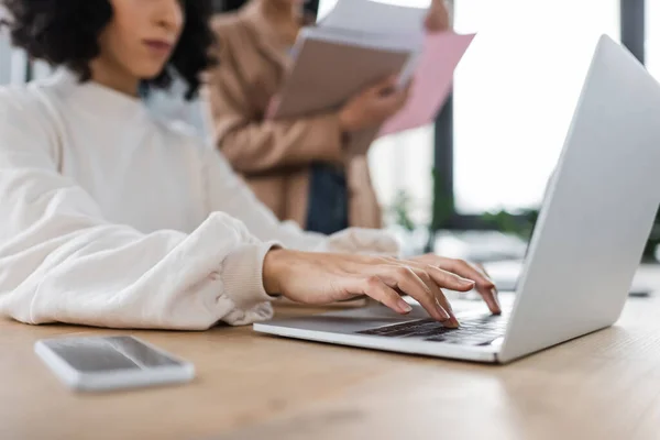 Cropped view of muslim businesswoman using laptop near blurred colleague in office - foto de stock