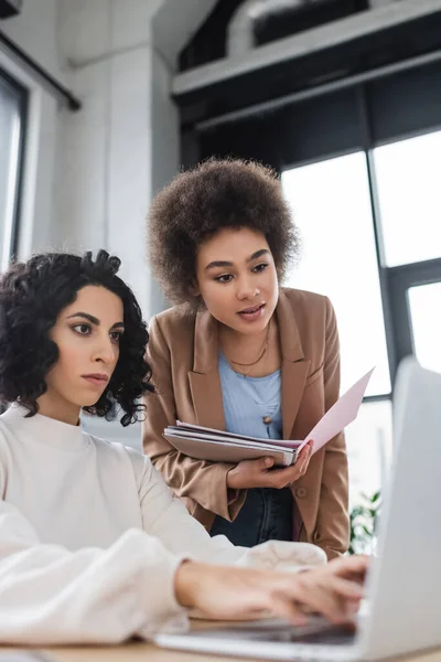 Donna d'affari afroamericana in possesso di cartella di carta vicino collega arabo utilizzando il computer portatile — Foto stock