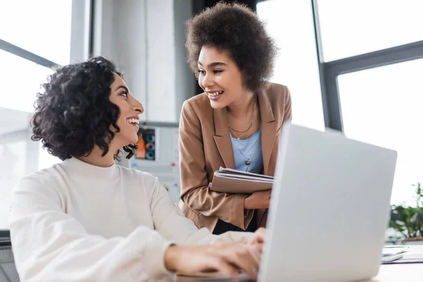 Lächelnde multiethnische Geschäftsfrauen reden in der Nähe von Laptop im Büro — Stockfoto