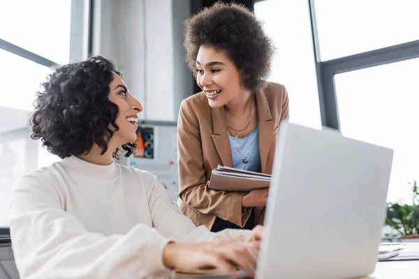 Souriantes femmes d'affaires multiethniques parlant près d'un ordinateur portable au bureau — Photo de stock