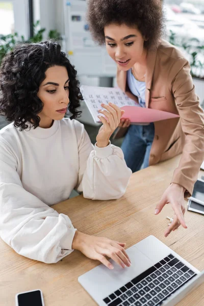 African american businesswoman pointing at laptop and holding paper folder near muslim colleague in office - foto de stock