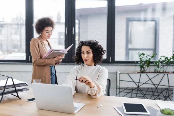 Smiling muslim businesswoman looking at laptop near devices and african american colleague with documents in office — Foto stock