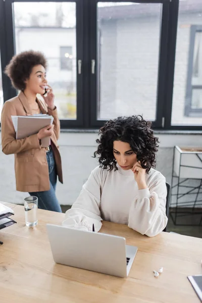 Arabian businesswoman using laptop while african american colleague holding papers and talking on smartphone — стокове фото