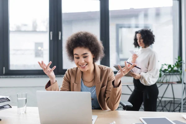 Fröhliche afrikanisch-amerikanische Geschäftsfrau mit Videochat auf Laptop in der Nähe verschwommener Kollegin im Büro — Stockfoto