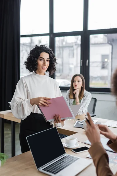 Smiling muslim businesswoman holding paper folder near devices and multiethnic colleagues in office — Fotografia de Stock