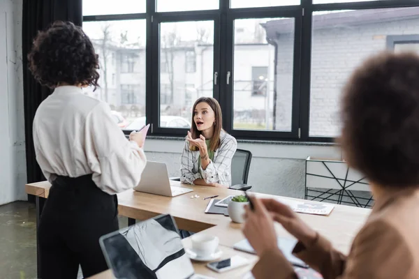 Geschäftsfrau im Gespräch mit arabischem Kollegen in der Nähe von Geräten und Papieren im Büro — Stockfoto