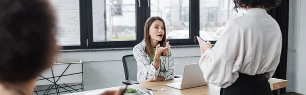 Young businesswoman talking to colleague near devices in office, banner — стоковое фото